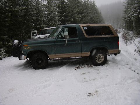 1991 Ford Bronco - A green and tan Ford Bronco sits in the snow on a wooded trail, with another vehicle partially visible behind it.