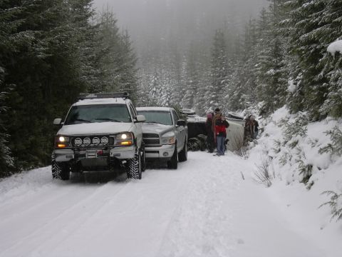1991 Ford Bronco - Two trucks assist a flipped vehicle on a snow-covered forest road.