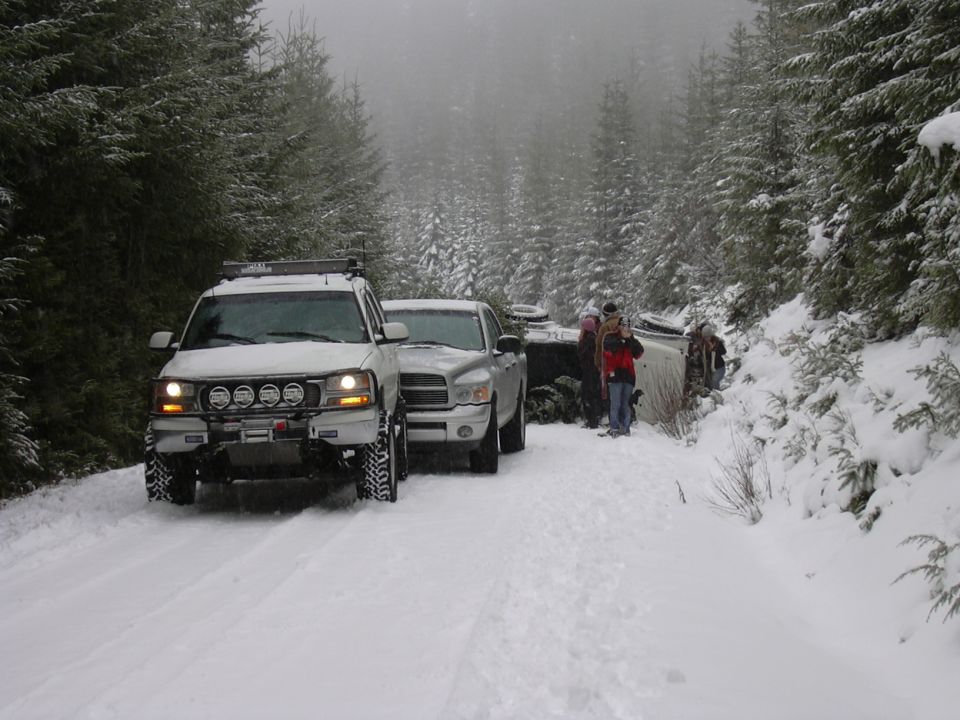 White truck and silver truck on snowy road next to flipped vehicle