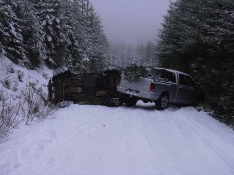 1991 Ford Bronco - A silver pickup truck carrying a Christmas tree is parked next to an overturned vehicle on a snow-covered road in a forest.