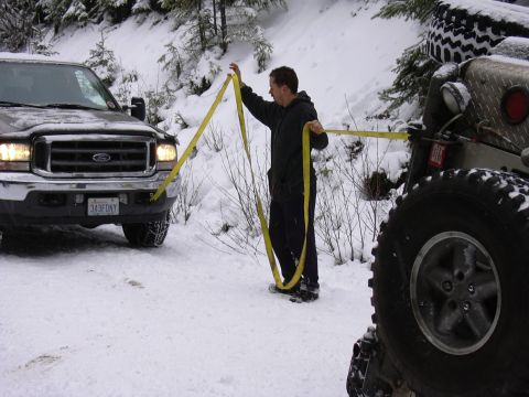 1991 Ford Bronco - A man is using a tow strap to connect a Ford truck to a Jeep in a snowy, wooded area.