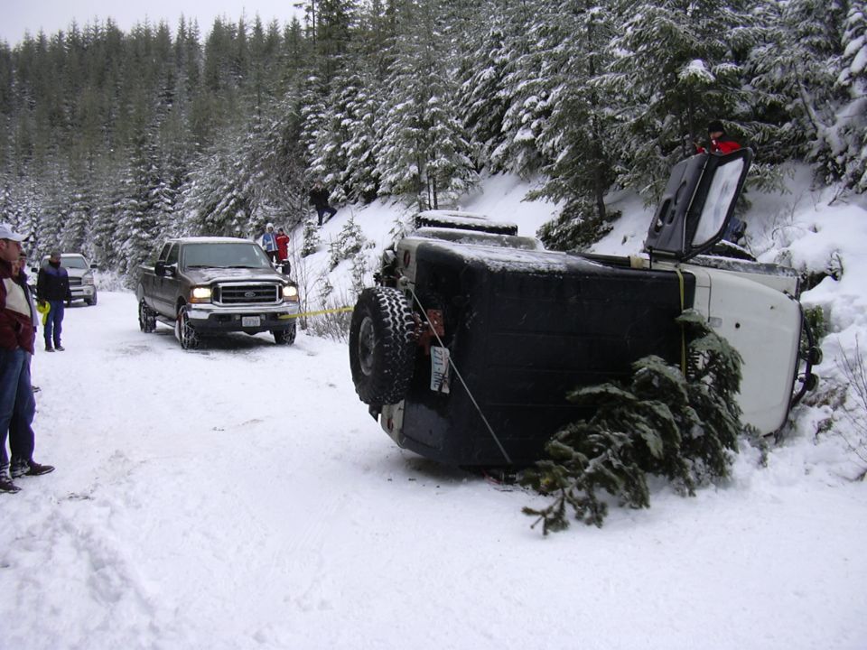 Overturned white SUV with a spare tire on the back, being towed by a brown pickup truck in a snowy forest