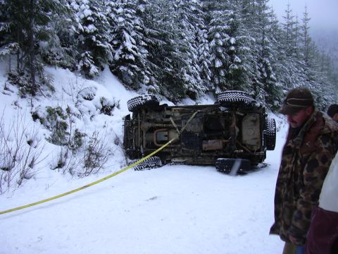 1991 Ford Bronco - An overturned vehicle lies on its side on a snow-covered road, attached to a yellow tow strap.