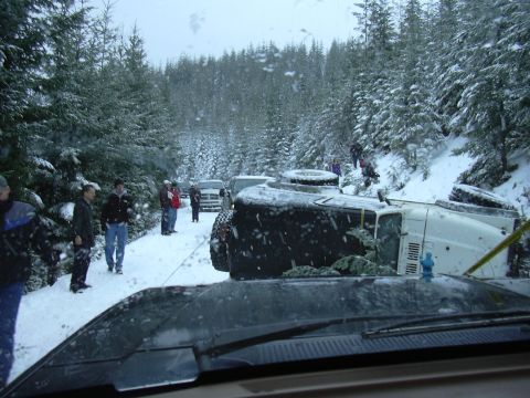 1991 Ford Bronco - Multiple vehicles and people are present on a snow-covered road next to a forest, with one vehicle overturned.