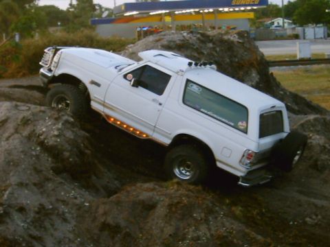 1995 Ford Bronco - White full-size Ford Bronco climbing over a dirt mound at dusk, viewed from the driver's rear three-quarter with the front wheels up on the…