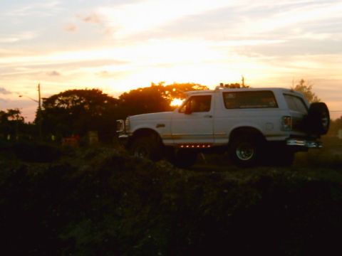 1995 Ford Bronco - White lifted Ford Bronco parked in profile on a dirt mound, backlit by a low orange sunset. The silhouette shows a roof light bar, rear-mou…