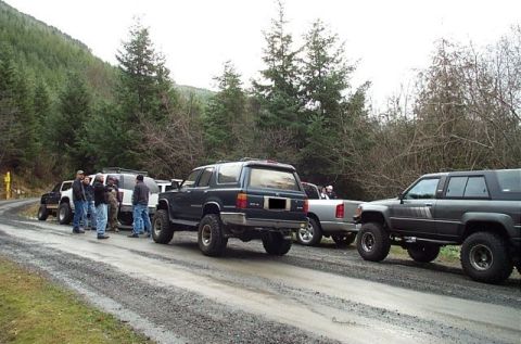 1995 Ford Bronco - A group of off-road vehicles are gathered on a dirt road, surrounded by a forest and a cloudy sky.