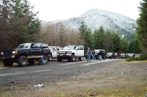 1995 Ford Bronco - A line of SUVs and trucks are parked on a gravel road with a snow-capped mountain in the background.
