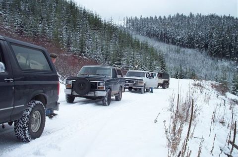 1995 Ford Bronco - Several SUVs drive in a line on a snow-covered road through a forest.