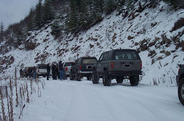 Several SUVs lined up on a snow-covered road next to a rocky, wooded slope