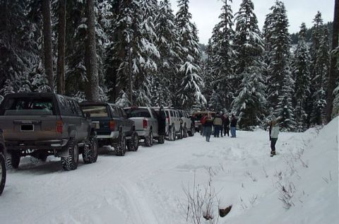1995 Ford Bronco - Several off-road vehicles are lined up on a snowy forest road, with a group of people gathered in the background.