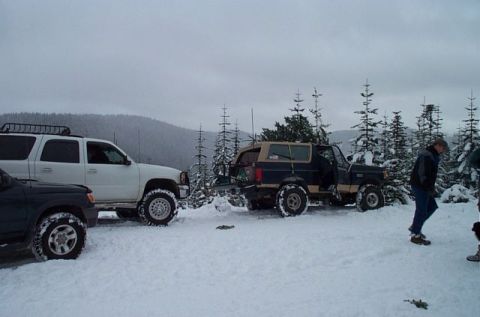 1995 Ford Bronco - A lifted dark blue and tan two-tone full-size Ford Bronco on oversized mud tires sits with its rear tailgate open in deep snow at a wooded…