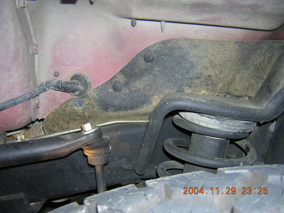 Red vehicle's suspension with coil spring, shock absorber, and control arm visible, covered in dirt