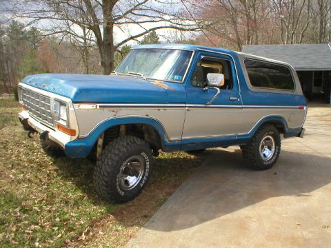 1978 Ford Bronco - Driver-side front three-quarter view of a blue and silver two-tone Ford Bronco parked on a leafy lawn beside a concrete driveway, showing s…