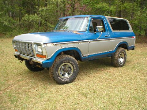 1978 Ford Bronco - 3/4 front view of a blue and white two-tone late-'70s Ford Bronco parked on grass in front of pine woods, with a removable white hardtop, c…