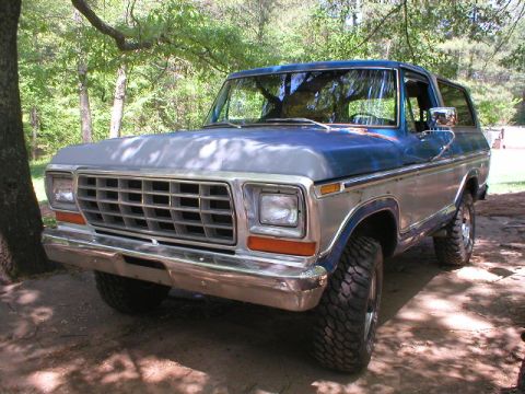 1978 Ford Bronco - Low 3/4 front view of a blue and silver two-tone late-'70s Ford Bronco parked in dappled shade on red dirt, showing the chrome egg-crate gr…