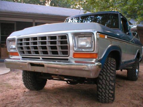 1978 Ford Bronco - Low front three-quarter shot of a blue and silver two-tone Ford Bronco parked on red dirt beside a house, emphasizing the wide egg-crate gr…