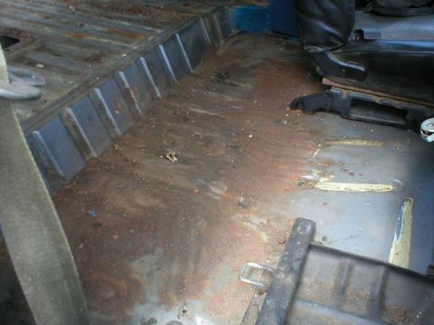 1978 Ford Bronco - Interior floor pan of a stripped truck or SUV with carpet and trim removed, showing surface rust across the ribbed rear floor and wheel-wel…