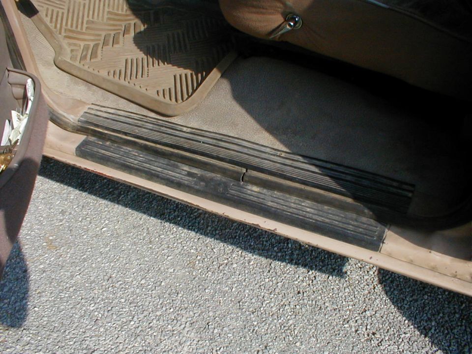 Worn rubber door sill step plate at an open vehicle door with a tan floor mat and gravel ground below