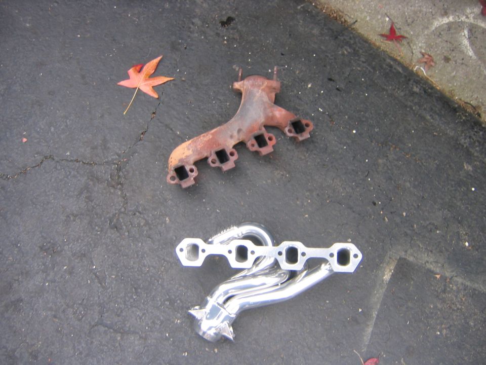 Rusty cast-iron exhaust manifold beside a chrome tubular header on pavement with fall leaves