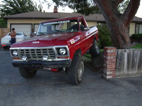 1978 Ford Bronco - Red pickup truck with lifted suspension is partially on a brick wall, with a white car in the background.