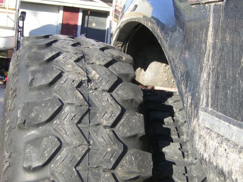 1995 Ford Bronco - Close-up comparison of two off-road tires leaning against a dirty blue truck body, showing an all-terrain tread pattern in the foreground n…