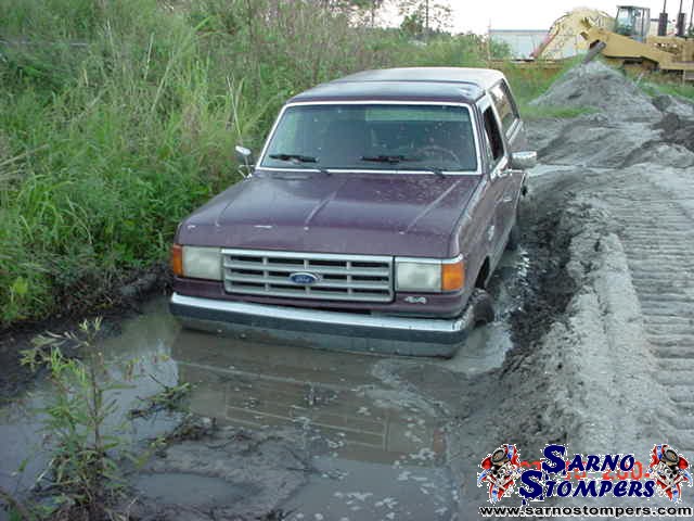 Maroon Ford Bronco stuck in deep mud and water next to a sand berm with an excavator behind