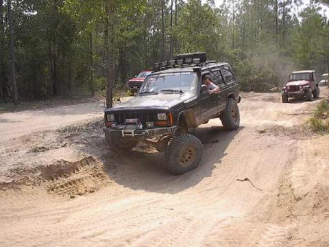 2000 Jeep Cherokee - A dark green Jeep Cherokee navigates a dusty dirt track with other vehicles in the background.