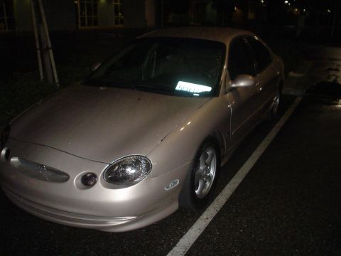 1993 Ford Taurus - A tan sedan is parked in a lot at night, showing its front 3/4 view with visible damage on the hood.