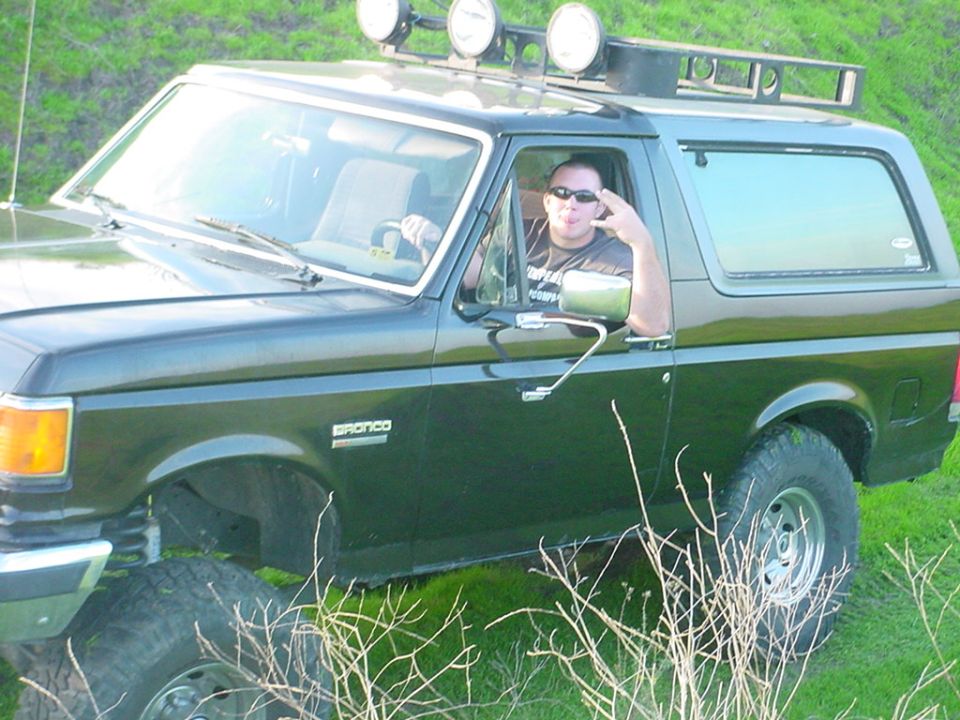 Man in sunglasses giving a peace sign from inside a dark green 1988 Ford Bronco with a roof rack and lights