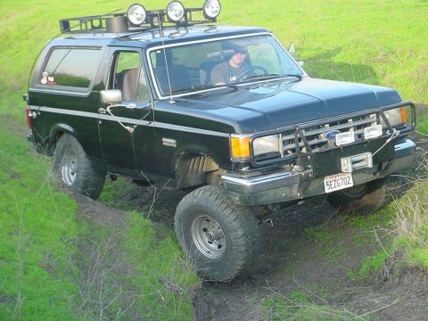 1988 Ford Bronco - This 1988 Ford Bronco is equipped with a roof rack and auxiliary lights, navigating a grassy, uneven terrain.