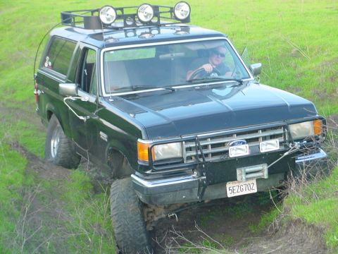 1988 Ford Bronco - A driver navigates a dark-colored Ford Bronco through a grassy, uneven terrain.