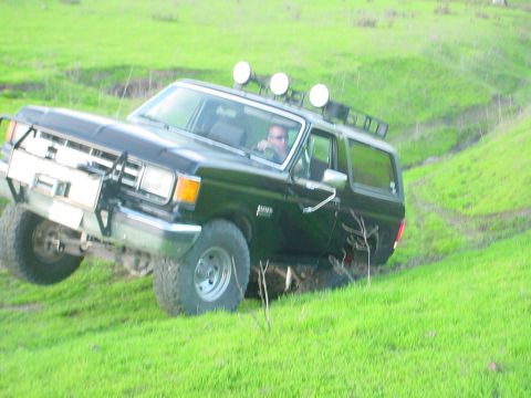 1988 Ford Bronco - A black Ford Bronco navigates a steep, grassy incline with auxiliary lights mounted on the roof.
