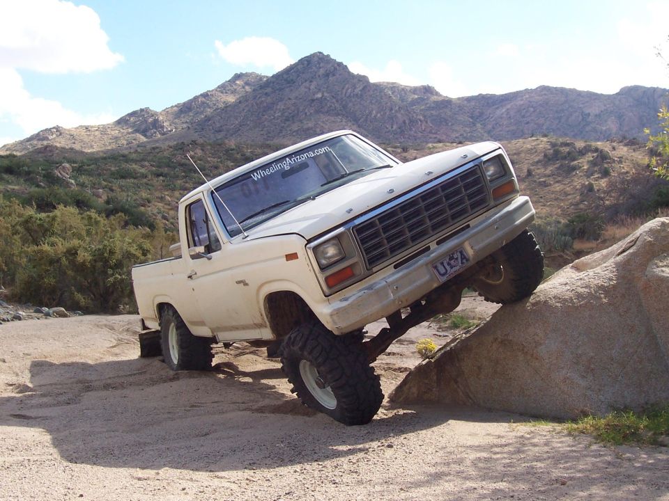 White lifted Ford pickup climbing a boulder off-road in desert terrain, front wheel on rock