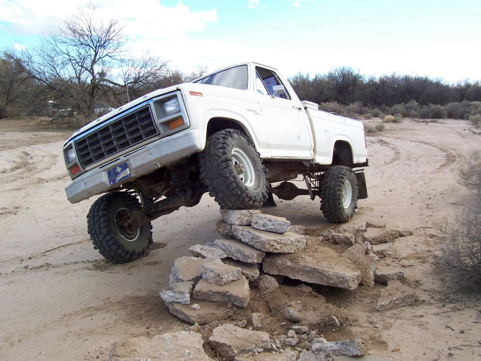 Lifted white Ford pickup on big mud tires flexing over broken concrete slabs in a desert area