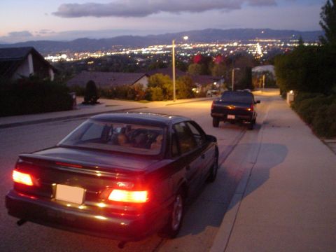 1993 Ford Taurus - A 1993 Ford Taurus is parked on a residential street at dusk, with a pickup truck behind it and a city skyline in the distance.