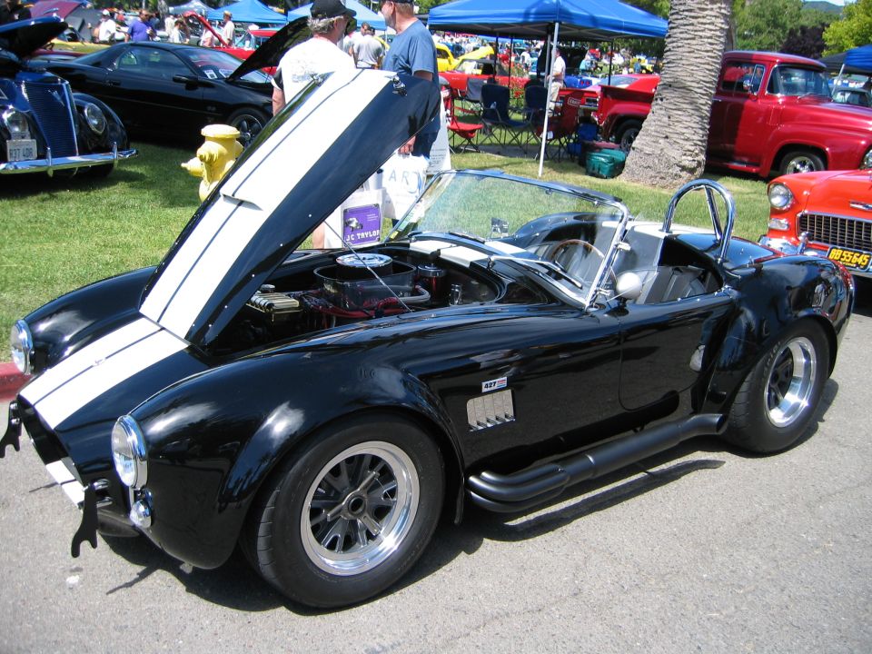 Black AC Cobra replica with hood open, showing engine. Other cars and tents in background