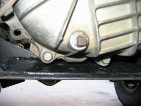1985 Ford Bronco - Underside view of a vehicle's transmission housing and skid plate, with visible bolts and a drain plug.