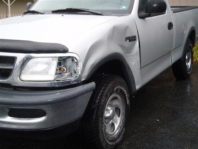 Silver Ford F-150 pickup with dented front fender and broken driver-side headlight, parked on wet pavement