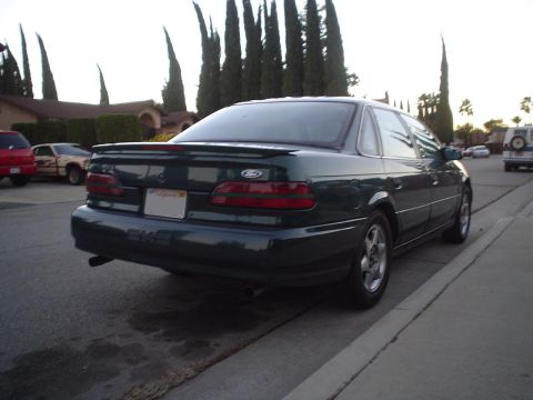 1993 Ford Taurus - Rear view of a dark green sedan parked on a residential street. Another vehicle and a van are in the background.