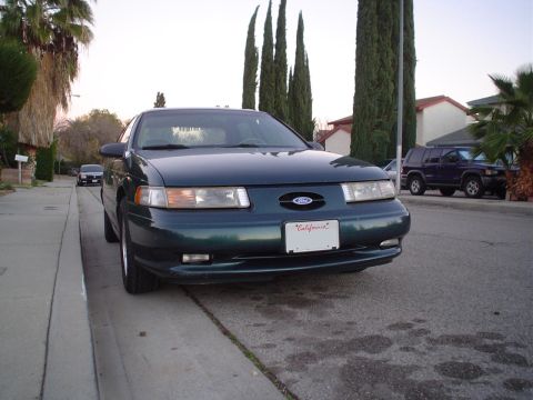 1993 Ford Taurus - A 1993 Ford Taurus is parked curbside on a residential street with trees and houses in the background.