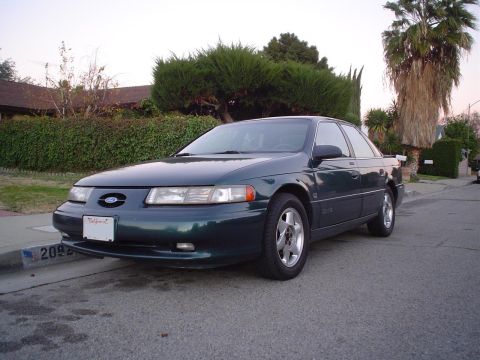 1993 Ford Taurus - A dark green 1993 Ford Taurus sedan is parked on the side of a street in front of a house and trees.
