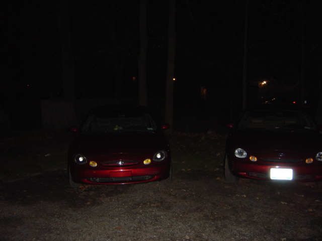 Two red sedans parked at night, front view. The car on the left has its headlights on