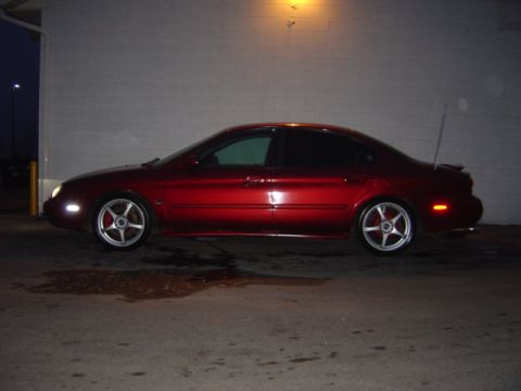 1998 Ford Taurus - A red 1998 Ford Taurus sedan is parked outside at night, showing its side profile with custom wheels.