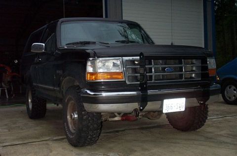 1995 Ford Bronco - Three-quarter front view of a lifted black Ford Bronco with removable hardtop parked on gravel outside a shop building, fitted with a black…