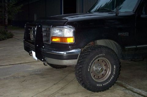 1995 Ford Bronco - Three-quarter front view of a black Ford Bronco at night with headlights on, wearing a black brush guard/winch bumper with D-ring, chrome b…