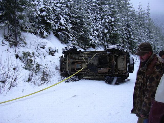 Towed vehicle flipped on its side in a snowy forest. A man in camouflage stands nearby