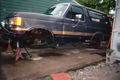 1990 Ford Bronco - black Ford Bronco SUV sits on jack stands with its wheels removed during suspension work in a driveway.
