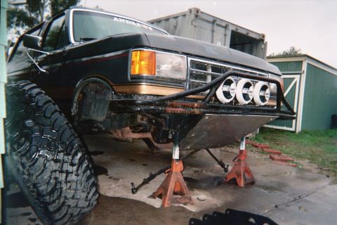 1990 Ford Bronco - Black and gold lifted Ford pickup raised on jack stands, fitted with a tubular front bumper and three round driving lights, in a shed.