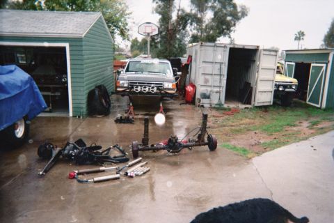 1990 Ford Bronco - truck raised on stands sits near garages and a shipping container while a removed axle and suspension parts lie on the wet pavement.